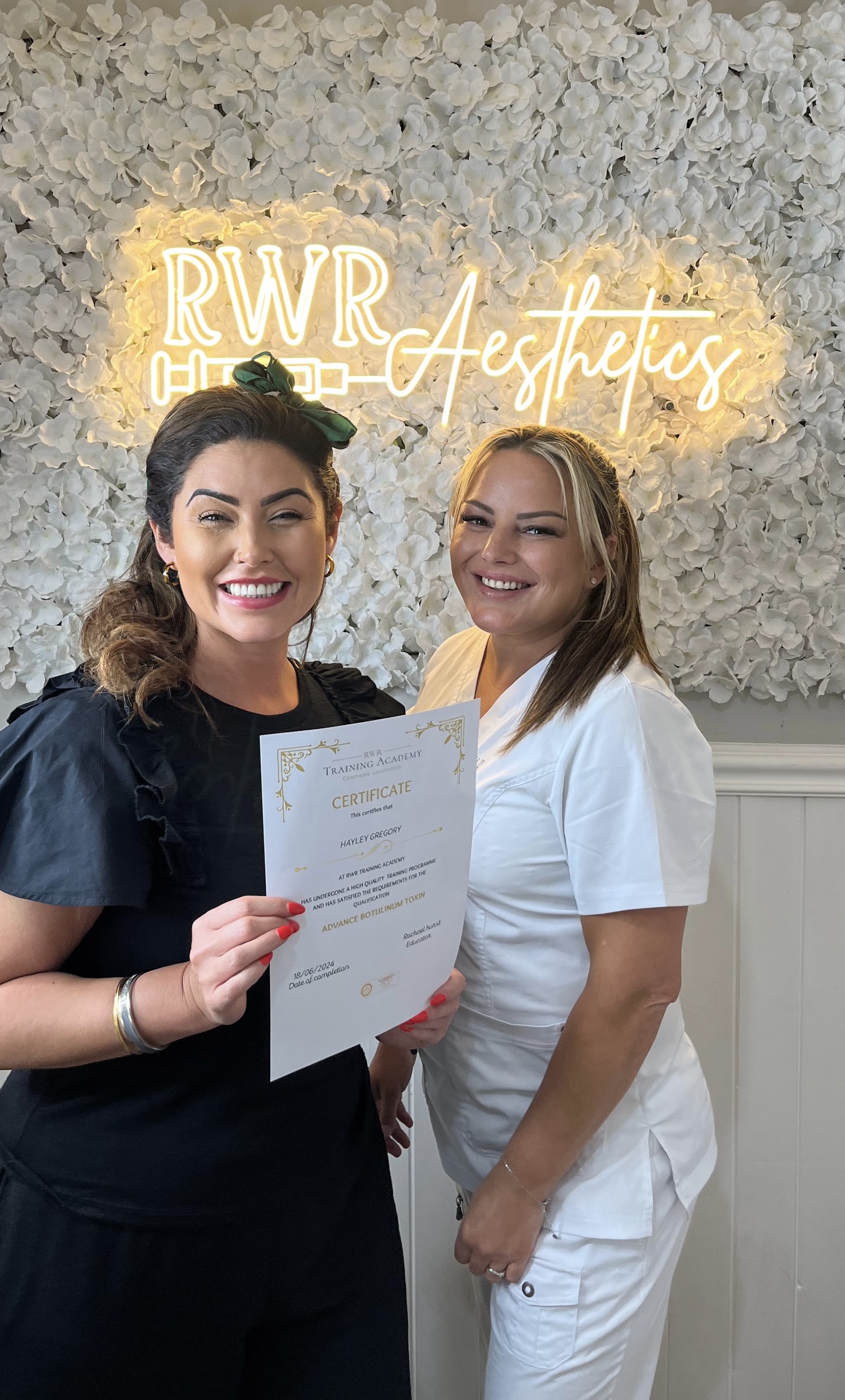 The RWR Aesthetics studio: a white flower wall with the warm neon RWR Aesthetics sign, two women smiling, one holding a certificate